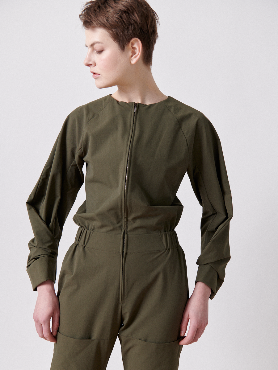 A person with short brown hair wears the Zero + Maria Cornejo Biker Jumpsuit – Final Sale in olive green, featuring an elastic banded waist for comfort. They look to the side with hands on hips against a plain light-colored background.