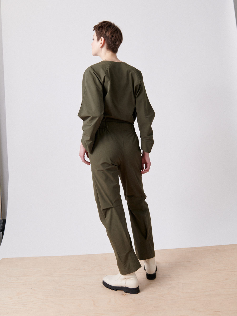 A person faces away against a plain white background, wearing the olive green Biker Jumpsuit from Zero + Maria Cornejo with an elastic waist and long sleeves, paired with light-colored boots. The wooden floor adds to the minimalist setting.