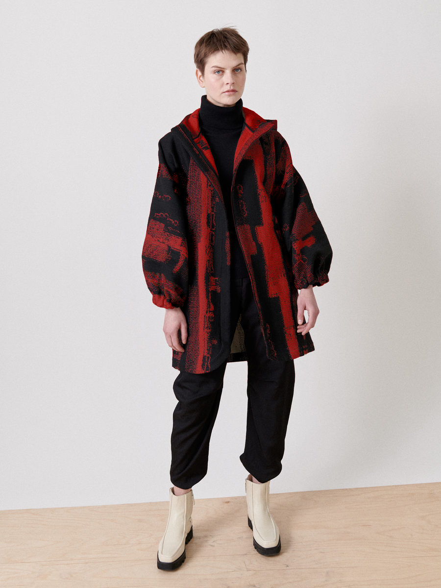 Dressed in a striking red and black jacquard Nio Coat from Zero + Maria Cornejo, a person with short hair confidently stands on a wooden floor. Their outfit is completed with light-colored boots against a plain background, featuring a black turtleneck and pants.