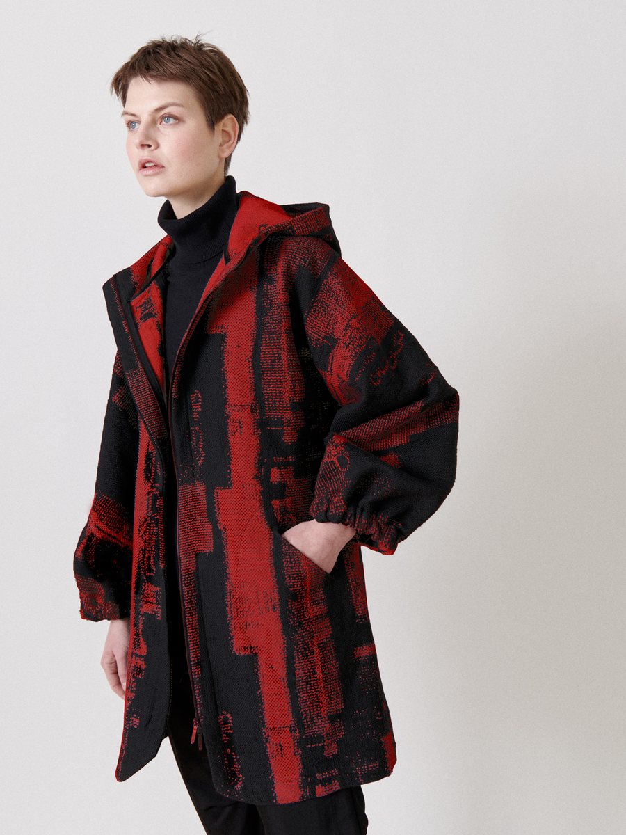 A person in a Zero + Maria Cornejo Nio Coat is wearing a black turtleneck under the jacquard hooded coat, featuring a striking red and black abstract pattern made from recycled fibers. They stand against a plain light background, looking sideways with hands in their pockets.