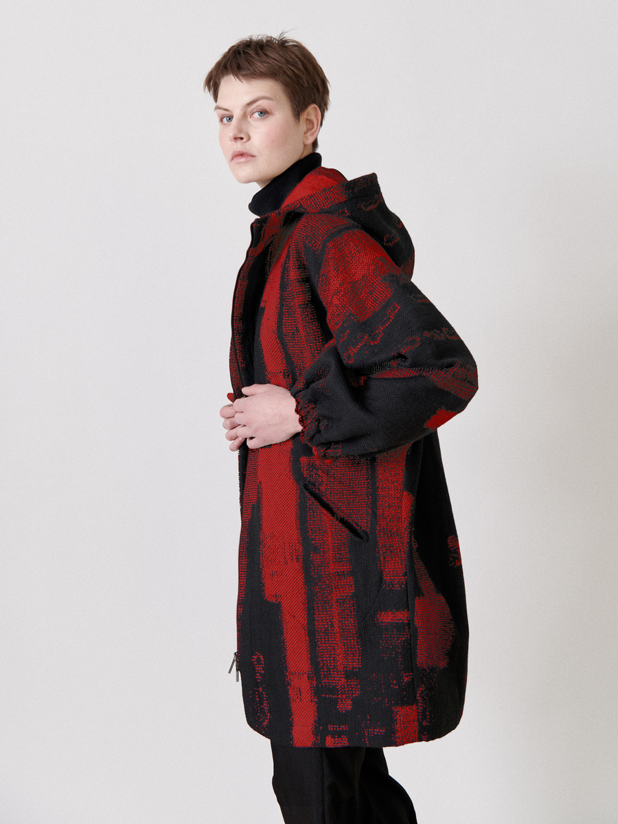 The person is wearing the Nio Coat by Zero + Maria Cornejo, a long-sleeved organic cotton coat with eye-catching red and black abstract patterns. They have short hair and are glancing over their shoulder against a plain white background.