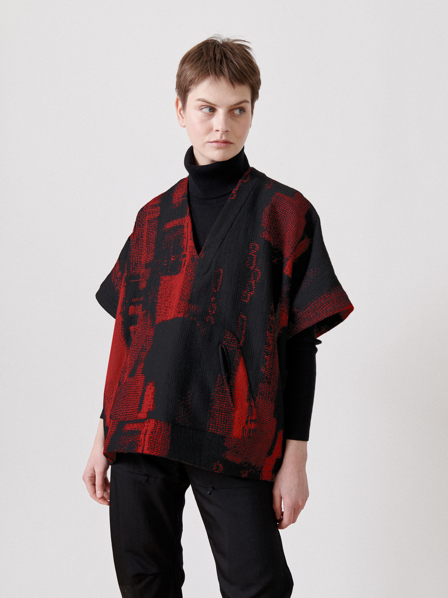 A person with short hair in a black turtleneck and a red/black patterned short-sleeve Elie Top from Zero + Maria Cornejo, made of recycled polyester, stands against a white background, looking to the side.