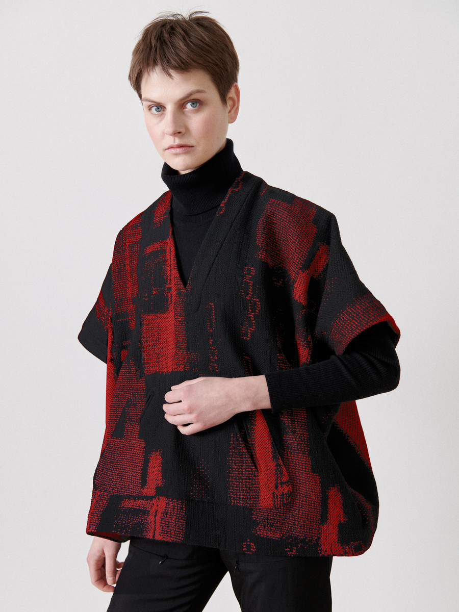 A person with short hair models the Elie Top from Zero + Maria Cornejo, a black turtleneck paired with a red and black jacquard poncho-like pullover. They pose against a plain background, one hand resting on their chest.