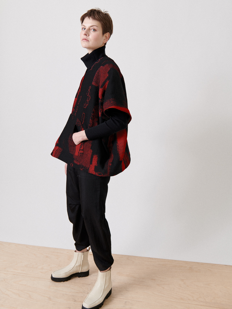 An individual with short hair, dressed in the Elie Top - Final Sale by Zero + Maria Cornejo, black pants, and beige ankle boots, stands in a minimalist indoor space featuring a light wood floor and plain white backdrop.