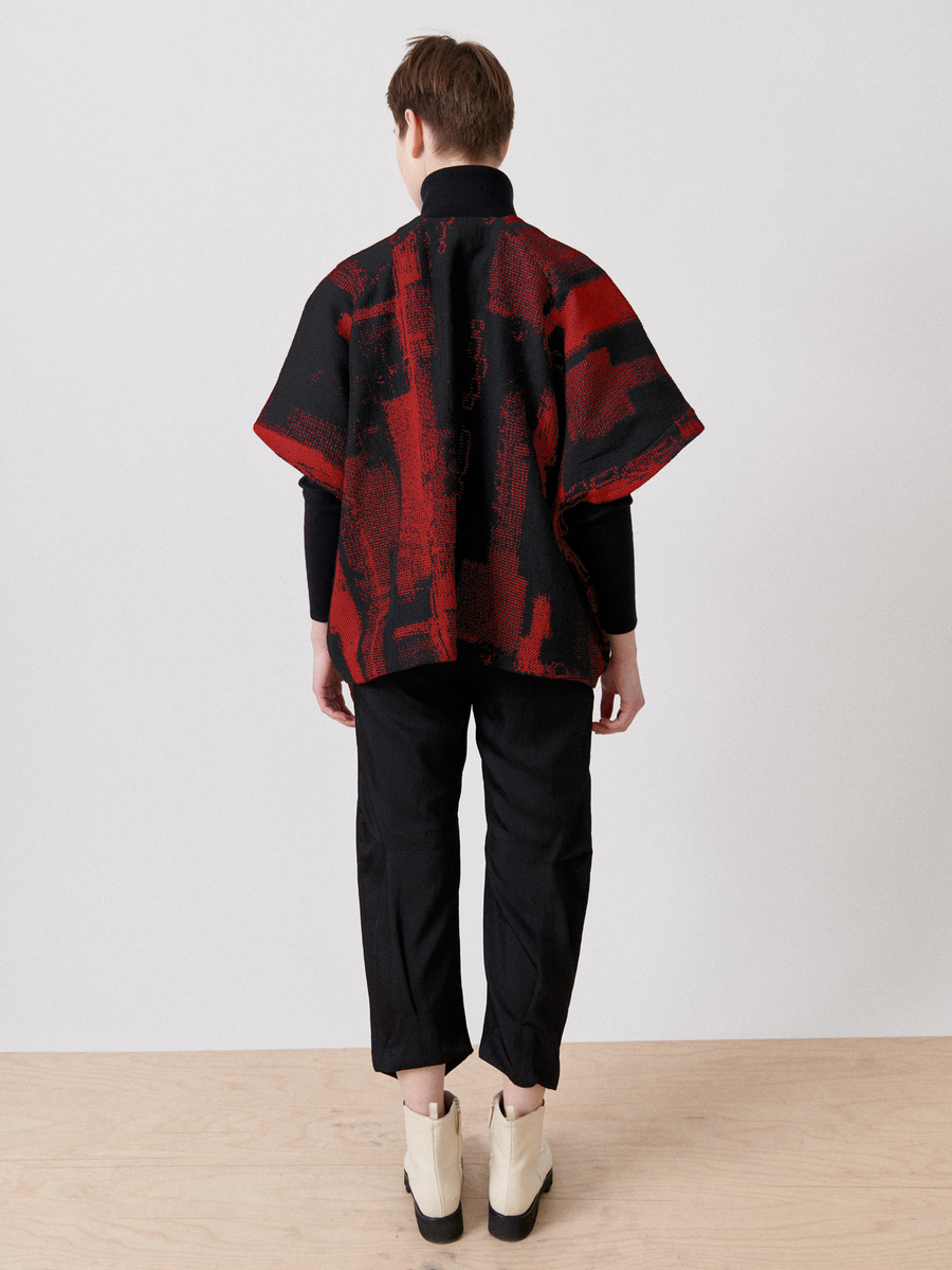 A person with their back to the camera wears an Elie Top - Final Sale, a red and black jacquard pullover from Zero + Maria Cornejo, paired with black pants and light-colored shoes. A simple light gray wall and wooden floor form the understated elegant backdrop.