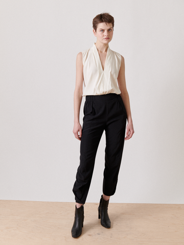 A person with short hair is wearing a sleeveless, white, v-neck top and high-waisted, black Zero + Maria Cornejo Akeo Pant featuring an elastic banded waist. They have black boots on and are standing on a light-colored wooden floor against a plain white backdrop.
