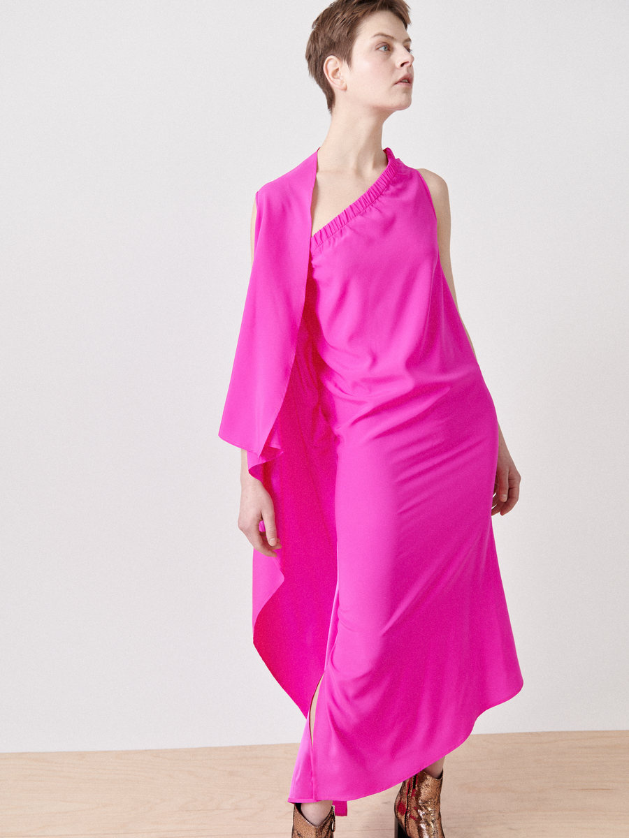 A person poses confidently in the One Chloe Dress by Zero + Maria Cornejo, showcasing its vibrant pink, silk charmeuse design with an asymmetrical neckline. Paired with patterned shoes, the outfit's bold color is highlighted against a plain background.