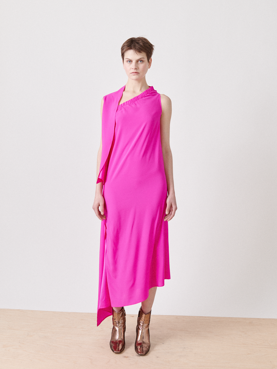 On a light wooden floor, someone wears the One Chloe Dress by Zero + Maria Cornejo in bright pink silk charmeuse featuring an asymmetrical neckline, paired with metallic high-heeled shoes. The plain white background highlights the clean, minimalist look.