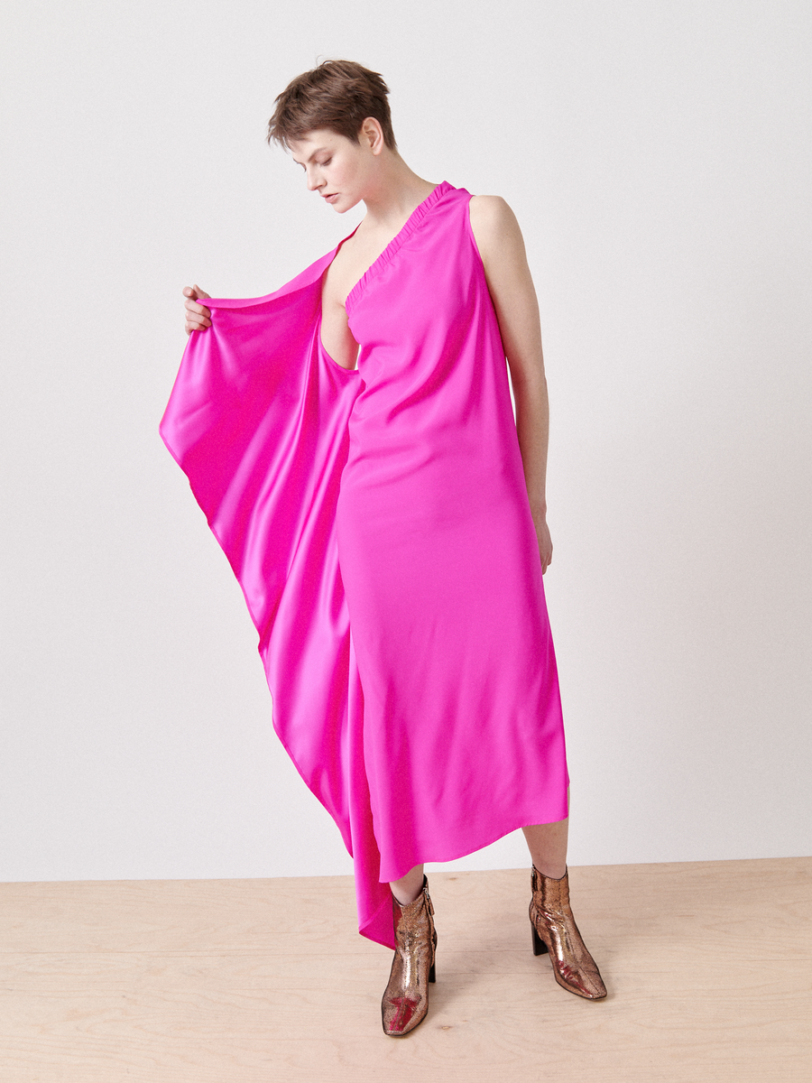 A person confidently stands on a light wooden floor wearing the One Chloe Dress, a bright pink silk charmeise with an asymmetrical neckline and flowing cape design. Their short hair complements the look, while shiny metallic boots shine against the plain white background. Brand: Zero + Maria Cornejo.