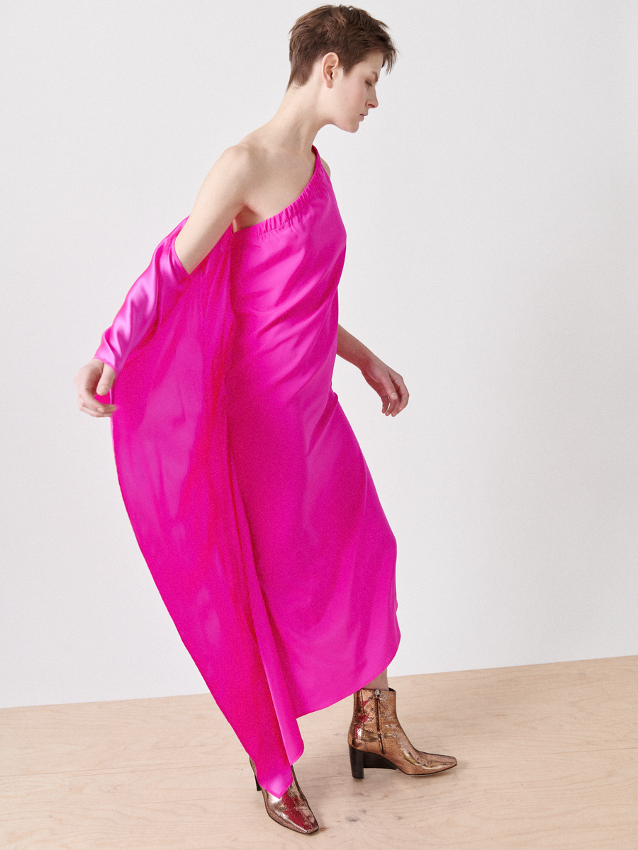 A person in the One Chloe Dress by Zero + Maria Cornejo, crafted in vibrant pink silk charmeuse with an asymmetrical neckline, pairs it with gold-patterned boots. Against a simple backdrop, their short hair complements the ensemble for a modern, stylish look.