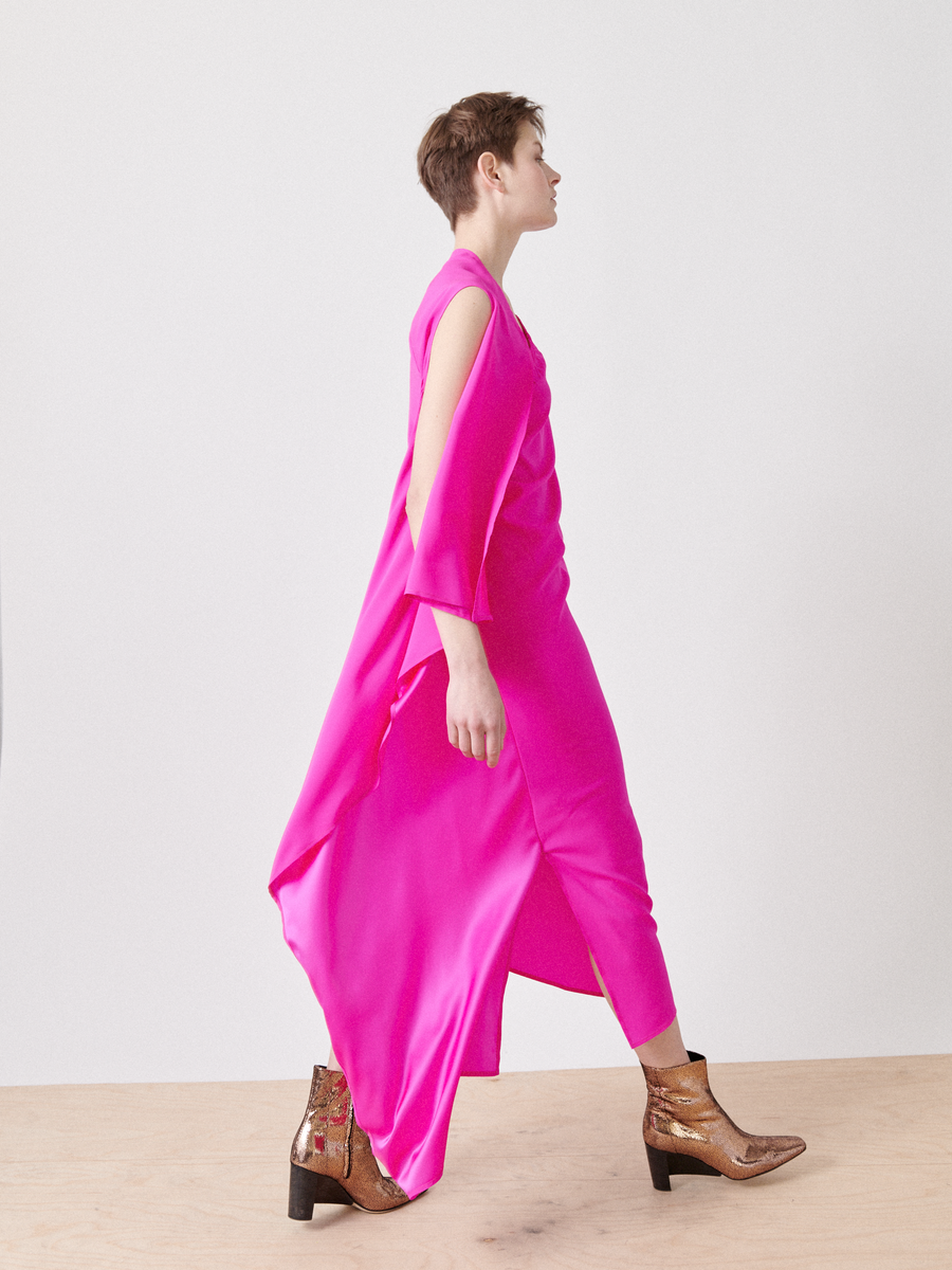 A person walks confidently on a wooden floor wearing the bright pink One Chloe Dress from Zero + Maria Cornejo, featuring side slits and an asymmetrical neckline. Metallic patterned ankle boots complete the look against a plain white background.