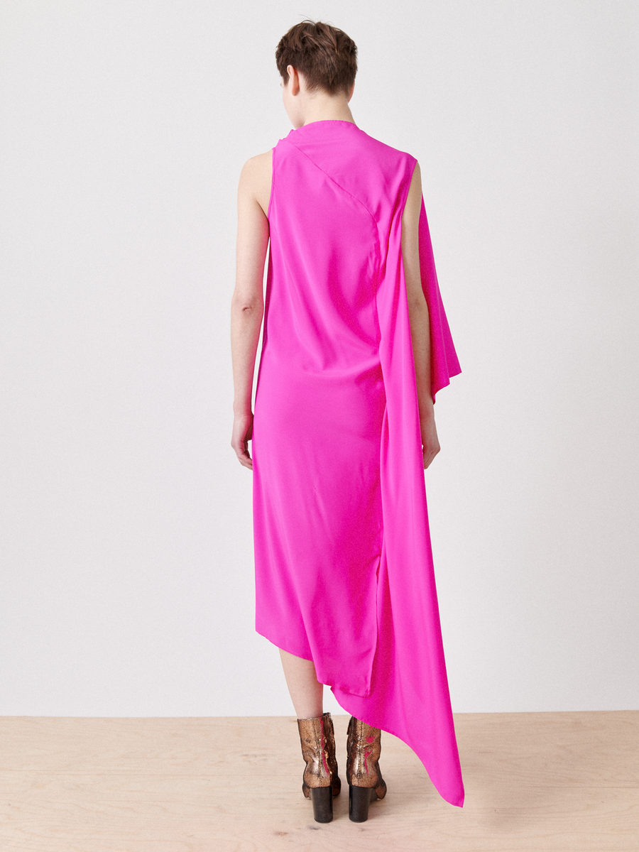 A person faces a white wall, wearing the One Chloe Dress by Zero + Maria Cornejo. It's a bright pink silk charmeuse dress with an asymmetrical neckline and a long draping piece on one side, paired with brown high-heeled boots. The floor is light wood.