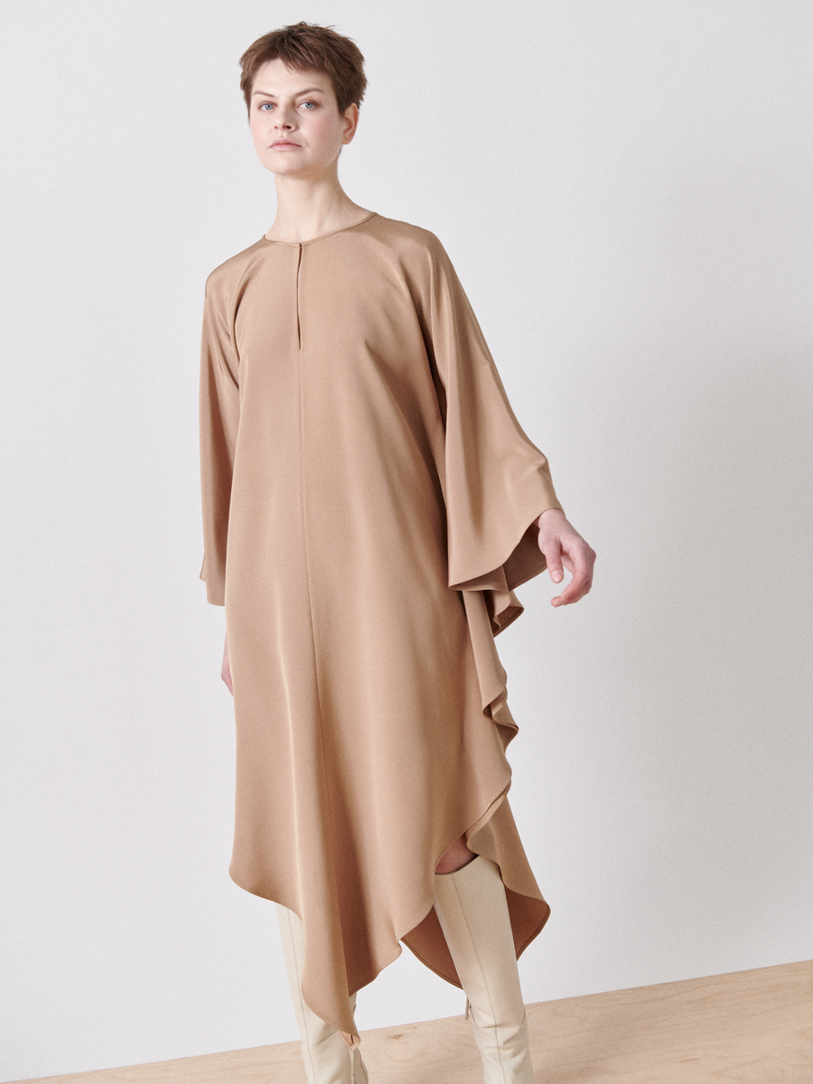 The Triangle Caftan by Zero + Maria Cornejo features a loose, camel-colored design with wide sleeves and a high-low hem against a plain background, showcasing an elegant draped silhouette made from responsible textiles.