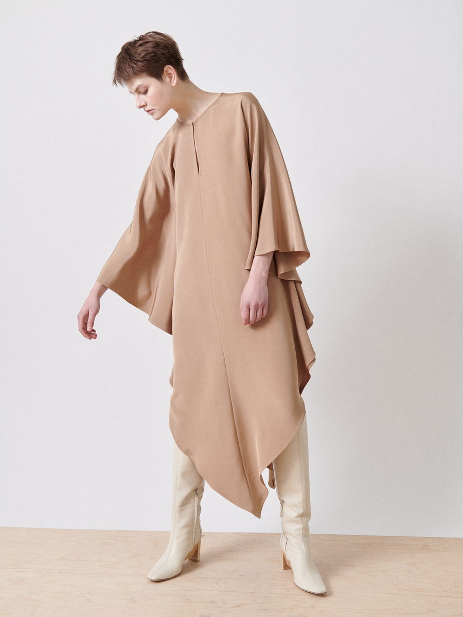 A person with short hair wears a Triangle Caftan from Zero + Maria Cornejo and tall off-white boots. The loose, tan silk dress drapes gracefully as they stand on a wooden floor with a neutral background, slightly leaning forward with arms relaxed.