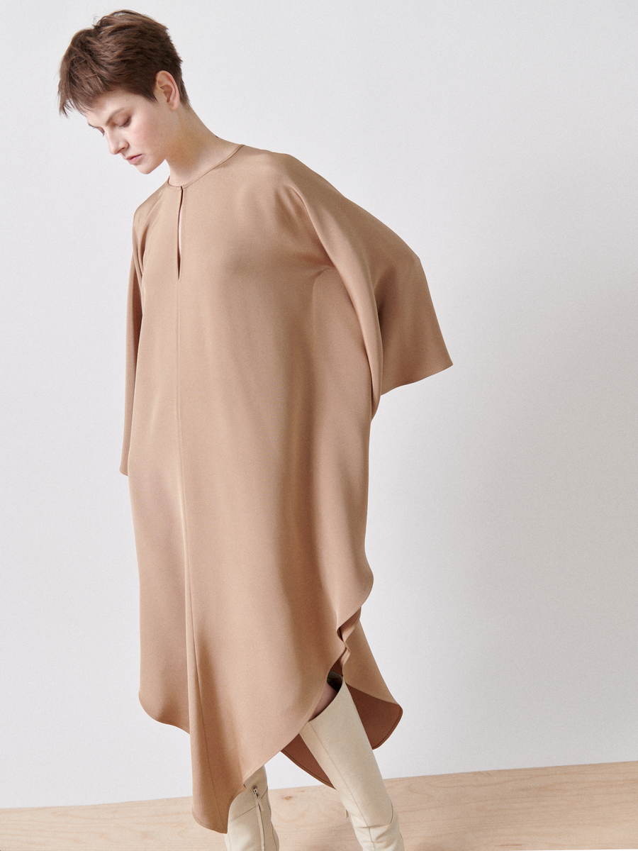 A person in a Triangle Caftan by Zero + Maria Cornejo stands against a plain white background, looking downward. They wear knee-high beige boots, creating a minimalist and elegant aesthetic with a beautifully draped silhouette.