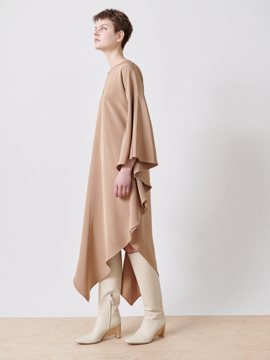 A person poses against a white backdrop in Zero + Maria Cornejo's Triangle Caftan, a loose tan silk dress with an asymmetrical hemline and wide sleeves. Paired with knee-high cream boots and short hair, the look is chic and crafted from responsible textiles.