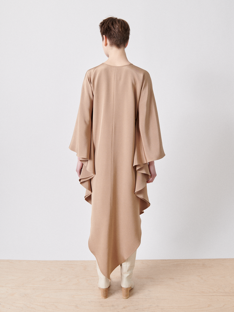 Facing away, someone wears the Triangle Caftan by Zero + Maria Cornejo, a long flowing beige garment with a draped silhouette and cascading fabric. Paired with light-colored pants and wooden shoes, it suggests responsible textiles against a plain white wall and light wooden floor.