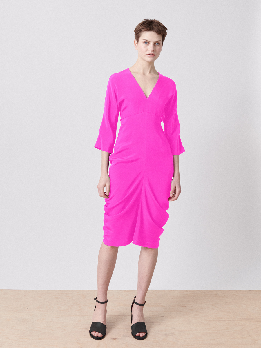 On a wooden floor against a white background, a person elegantly wears the Shae Dress in bright pink silk from Zero + Maria Cornejo, featuring an empire waist and three-quarter sleeves. The knee-length dress pairs perfectly with their black high-heeled sandals.