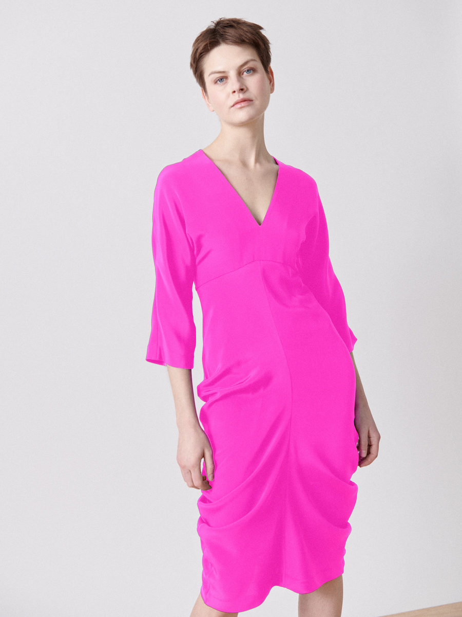 A person stands against a plain light gray background, wearing the Shae Dress by Zero + Maria Cornejo. The bright pink silk garment falls to the knee and has three-quarter sleeves, a V-neckline, and an empire waist, adding elegance to its vivid style.