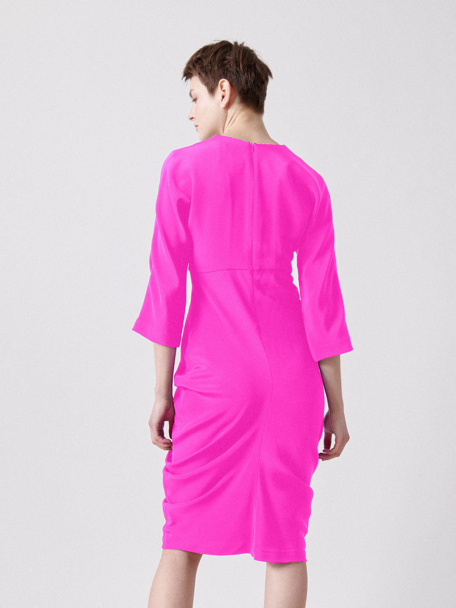 A person with short hair is wearing the Shae Dress - Final Sale by Zero + Maria Cornejo. The bright pink, silk dress has an empire waist and elbow-length sleeves. They face away from the camera in a neutral pose against a plain light gray background.