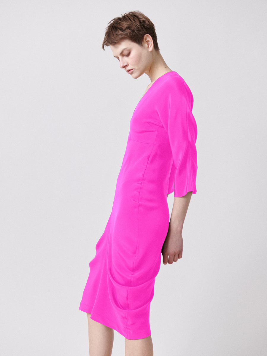 A short-haired individual in a knee-length, bright pink Shae Dress with an empire waist and three-quarter sleeves by Zero + Maria Cornejo stands against a plain white backdrop, gazing downwards to the left.