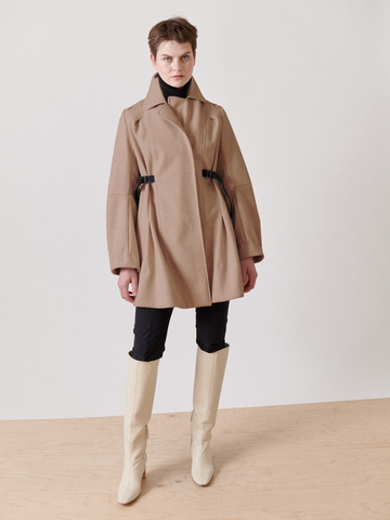 A person stands against a plain backdrop wearing the Leila Coat - Final Sale from Zero + Maria Cornejo, adorned with black accents, paired with black leggings and cream-colored knee-high boots. The tan wool coat's oversized collar adds elegance. They have short hair and face the camera.