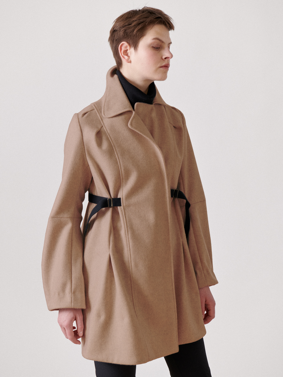 Someone wears the Leila Coat - Final Sale by Zero + Maria Cornejo, camel-colored and crafted from wool. It features black straps, an oversized collar, a tailored fit, unique sleeves, and a sleek grosgrain belt against a plain white background.