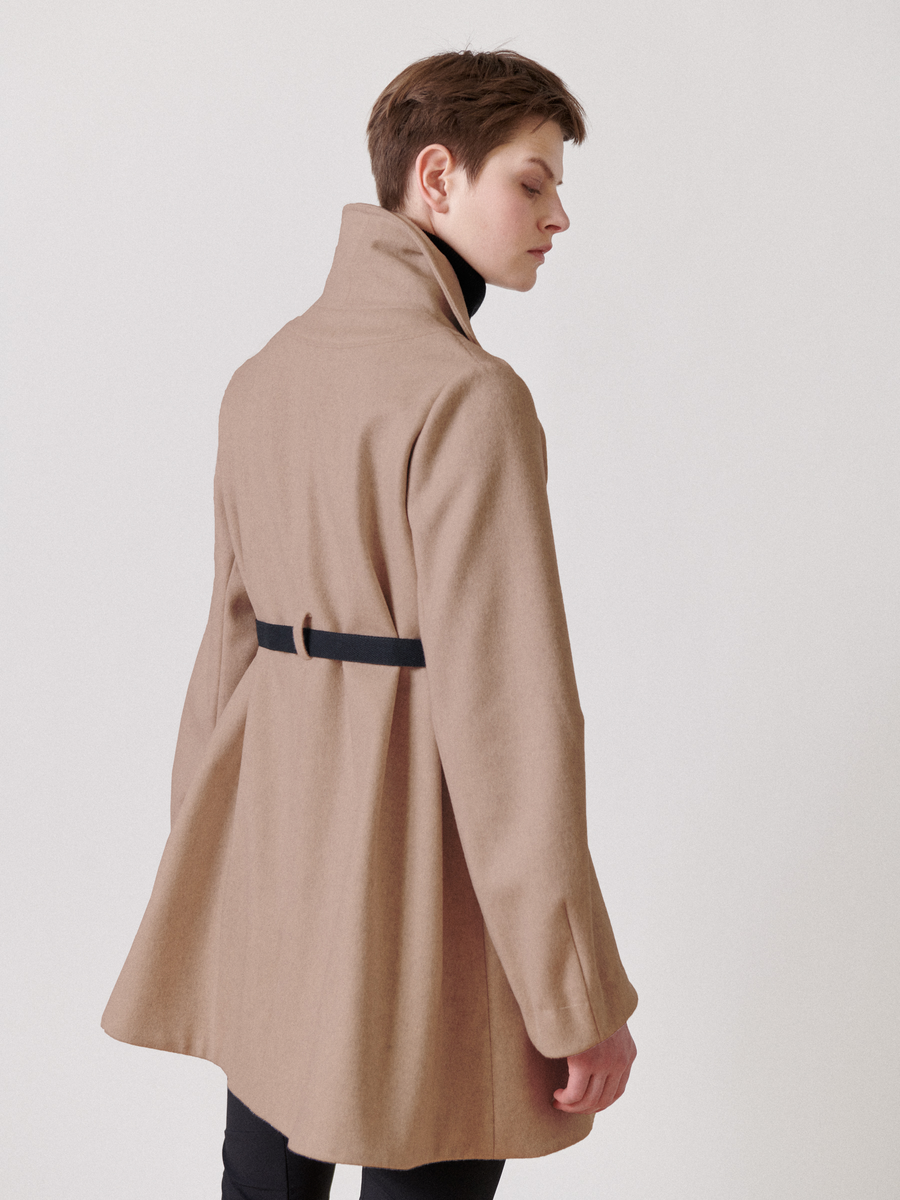 Against a plain white background, a person wears the Zero + Maria Cornejo Leila Coat - Final Sale, featuring a long beige wool design with an oversized collar and black grosgrain belt at the back, gazing thoughtfully to one side.