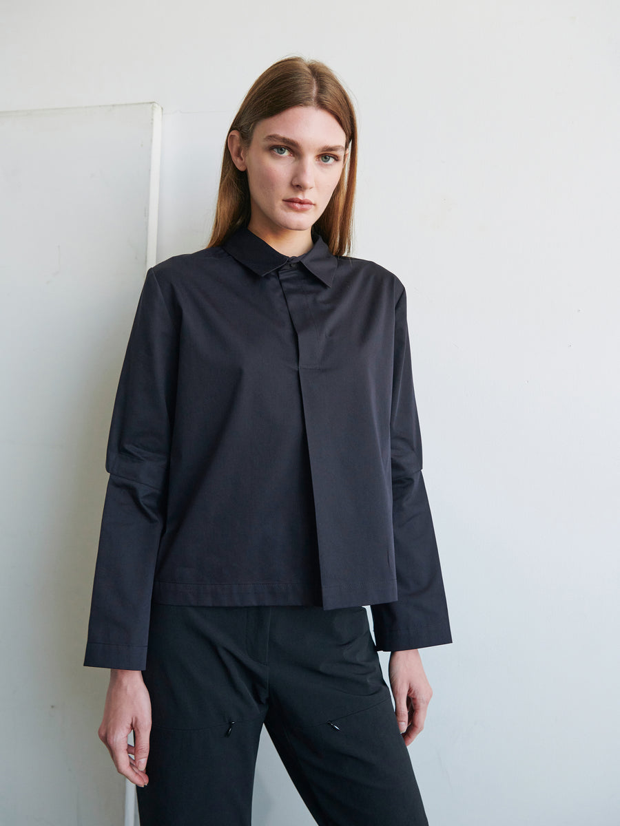 A person with straight brown hair stands against a white wall wearing the long-sleeved Biker Shirt from Zero + Maria Cornejo and matching dark pants. They are looking directly at the camera with a neutral expression.