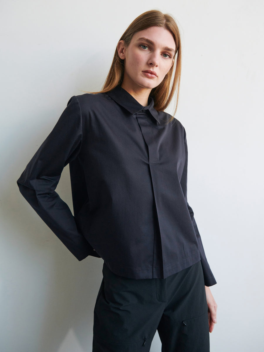 A person with long brown hair is wearing a black Biker Shirt by Zero + Maria Cornejo. The shirt features articulated sleeves and a stylish collar. They are standing against a plain, light-colored background, with one arm bent and resting on their hip while slightly leaning against the wall. Their expression is neutral.