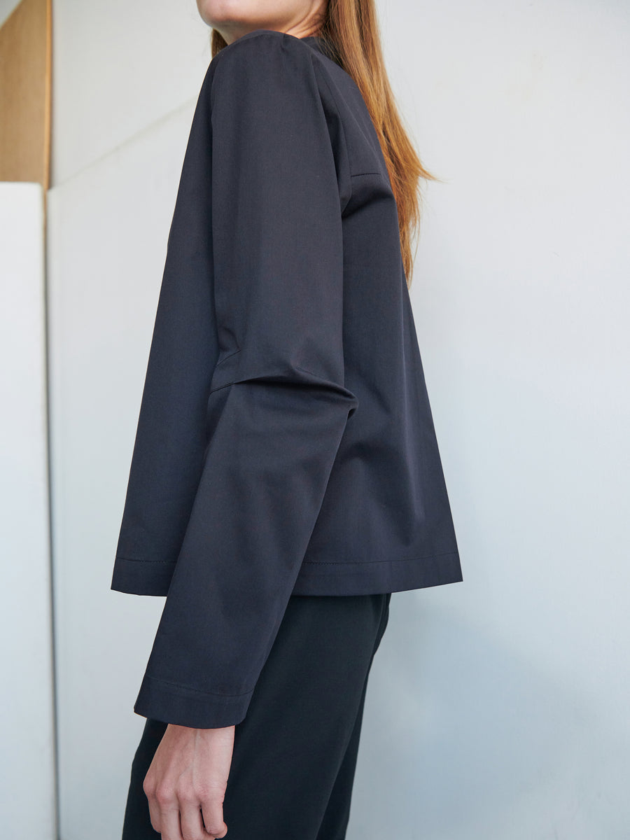 A person with long hair stands sideways facing left, wearing a dark Biker Shirt by Zero + Maria Cornejo and matching pants. The background is a minimalist, light-colored wall with a wooden panel partially visible.