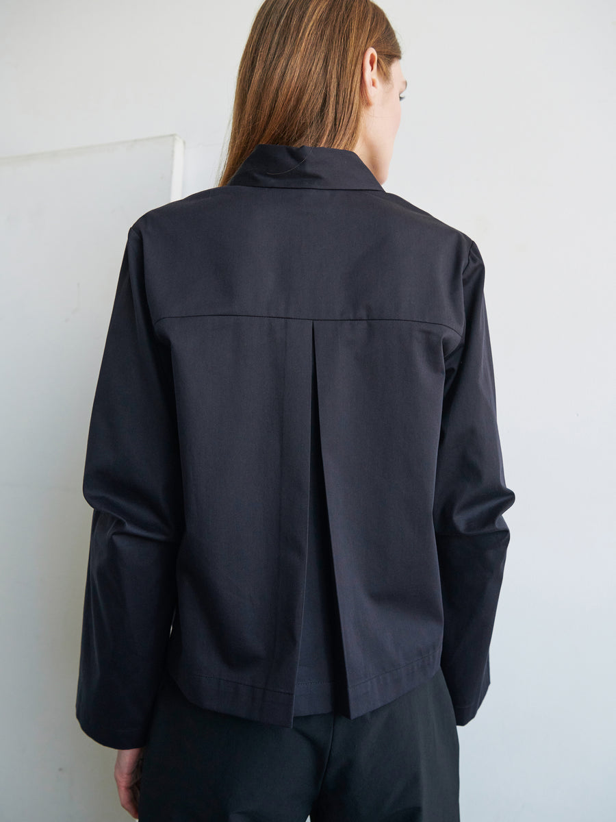 A person with long hair is standing with their back facing the camera, wearing a dark-colored Zero + Maria Cornejo Biker Shirt made from cotton broadcloth, which features a pleat in the middle of the back. The background is plain and light-colored.