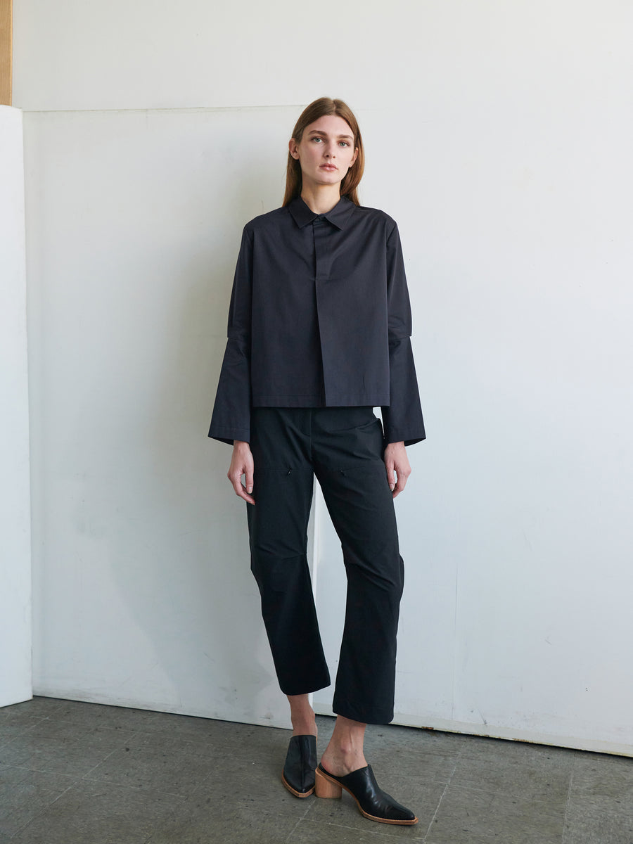 A person with long hair stands against a plain light-colored wall. They are wearing a black, loose-fitting cotton broadcloth Biker Shirt from Zero + Maria Cornejo, black cropped pants, and black slip-on shoes with a low heel. The floor is gray and the overall setting is minimalist.