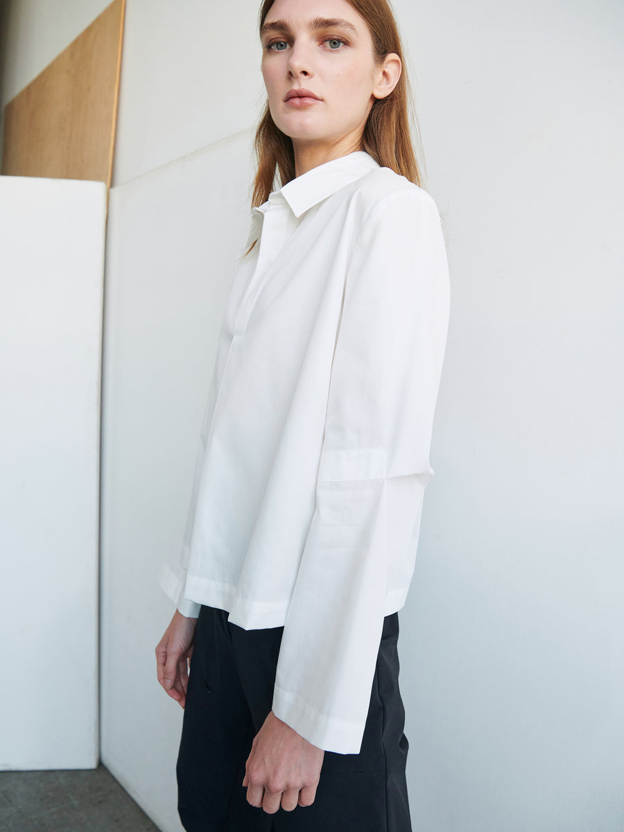 A person with long hair stands indoors wearing a loose, white Biker Shirt from Zero + Maria Cornejo and black pants. They are looking directly at the camera with a neutral expression, and the background includes white and beige walls.