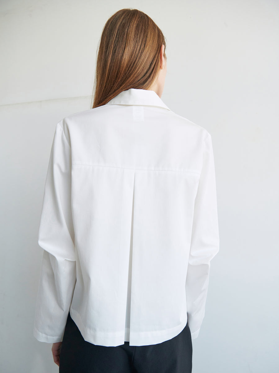 A person with long hair is seen from behind, wearing a white Biker Shirt made of cotton broadcloth, which features a pleat in the center of the back. The person also wears dark pants. The wall in the background is light-colored. This shirt is by Zero + Maria Cornejo.