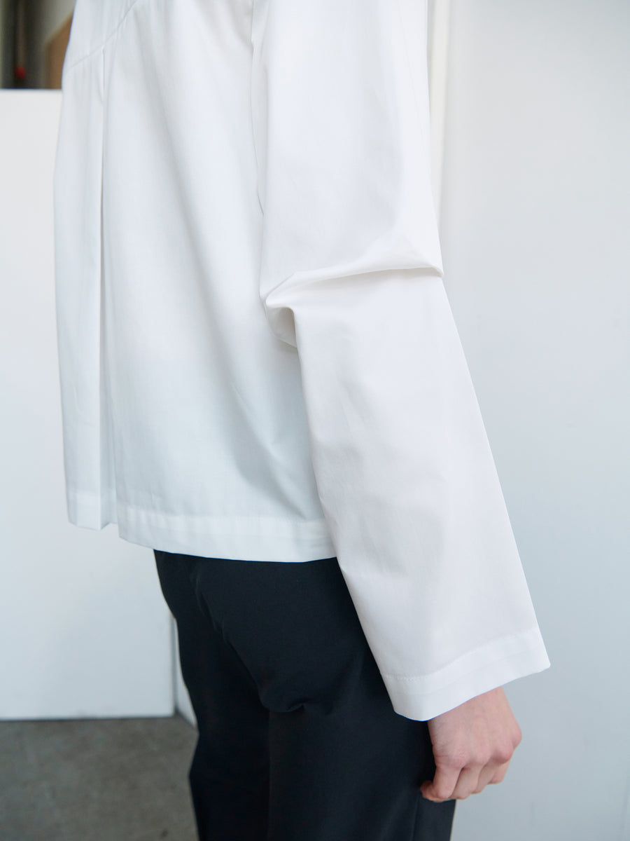 A person wearing a Zero + Maria Cornejo Biker Shirt and dark pants stands with their side facing the camera. The focus is on the torso, arm, and hand. The background is predominantly white with some grey flooring visible.