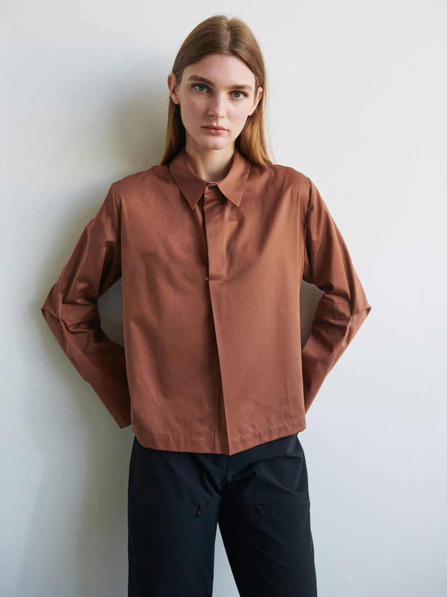 A person with long hair is standing against a plain white background. They are wearing the Biker Shirt by Zero + Maria Cornejo, featuring brown cotton broadcloth and articulated sleeves, paired with black pants. They have a neutral expression and their hands are resting on their hips.