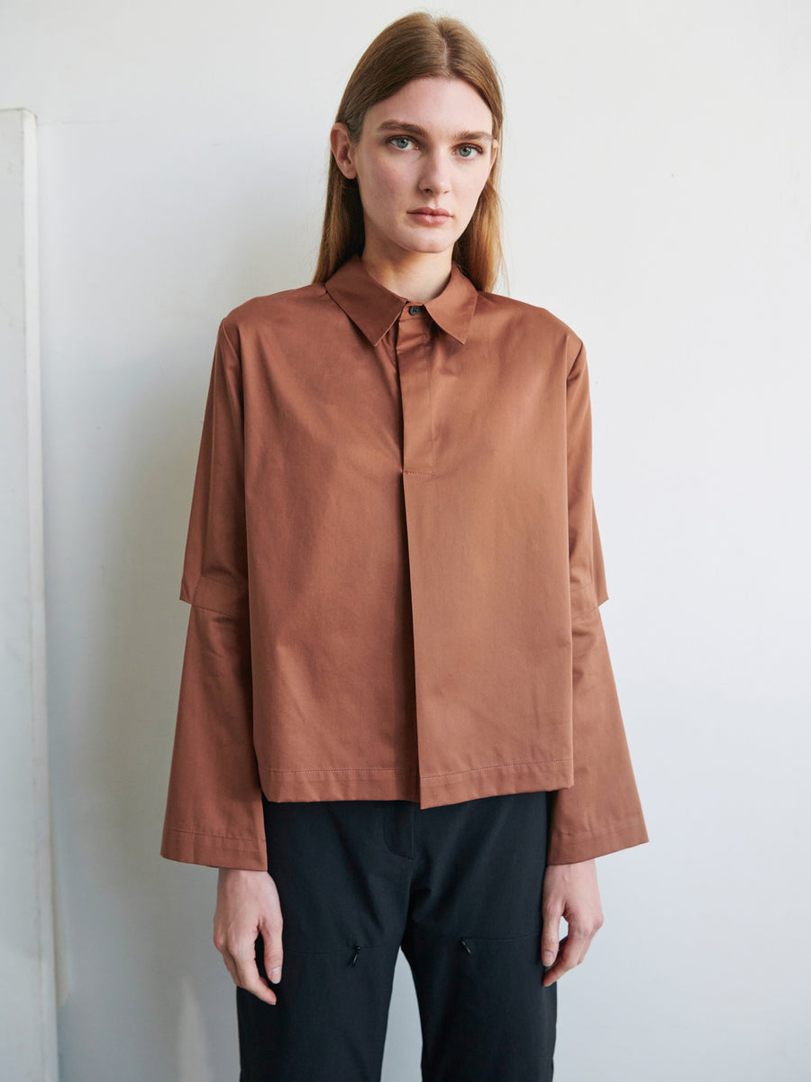 A person with long, light brown hair is standing against a white background. They are wearing a loose-fitting, long-sleeved Biker Shirt with articulated sleeves by Zero + Maria Cornejo and black pants. The person's expression is neutral.