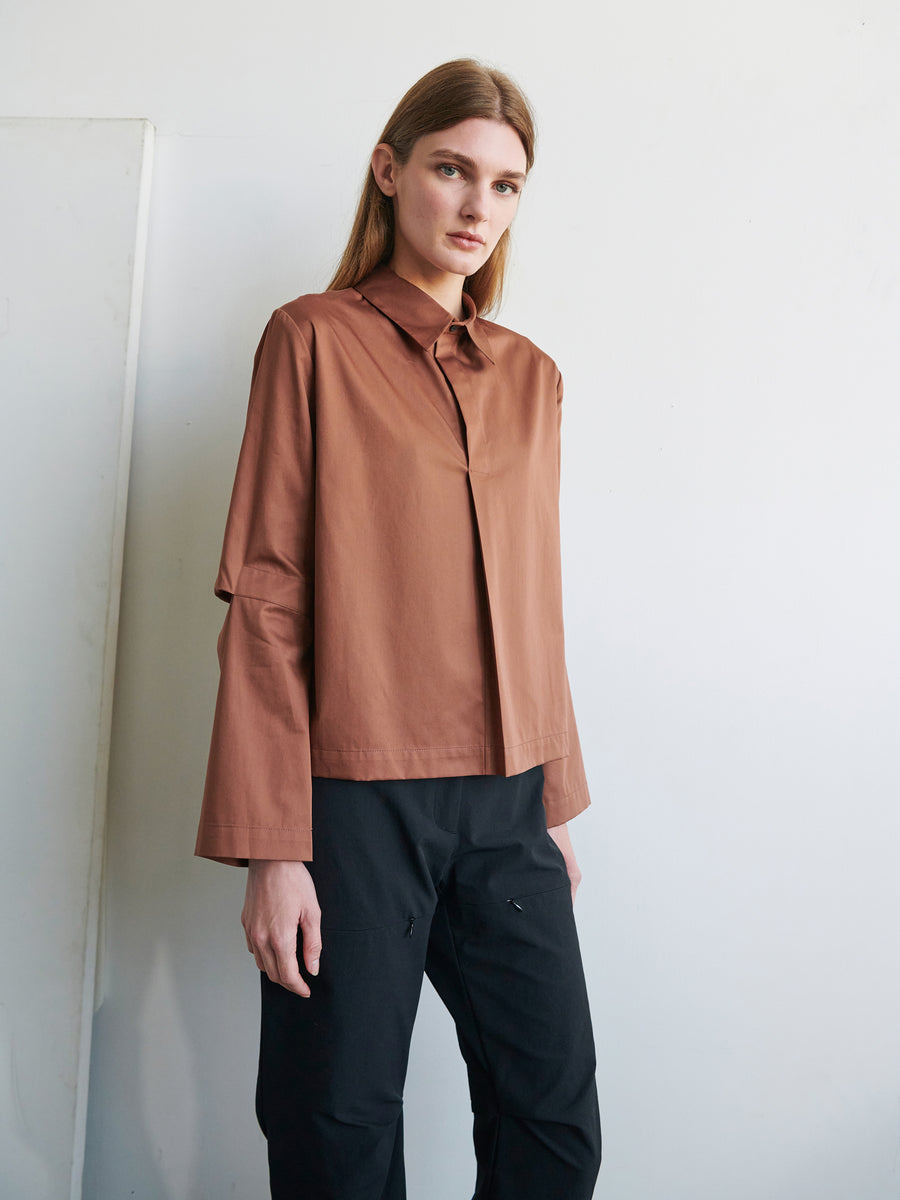 A person with long hair wearing a brown Biker Shirt made of cotton broadcloth by Zero + Maria Cornejo and black pants stands against a plain white background. The individual gazes directly at the camera with a neutral expression.