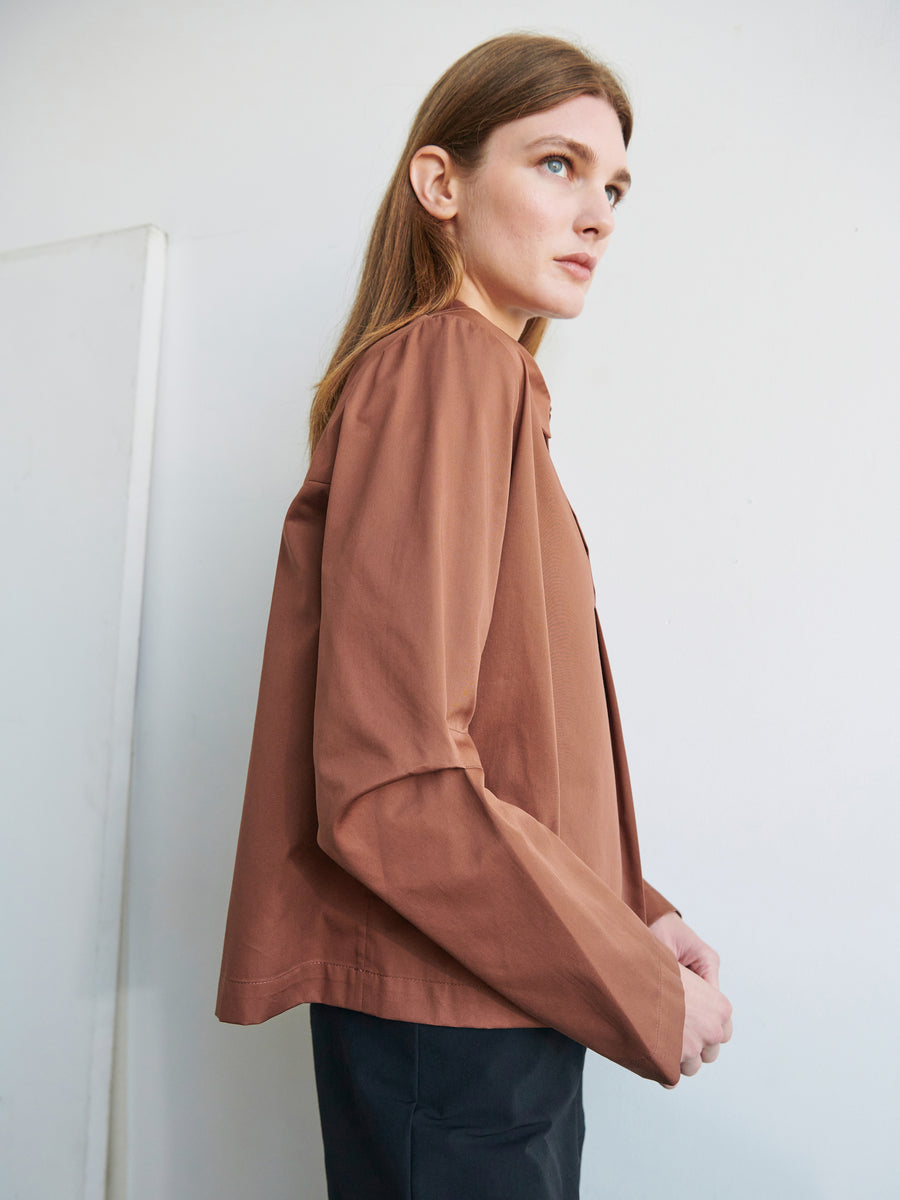 A person with long light brown hair stands against a plain background. They are wearing a loose-fitting Biker Shirt from Zero + Maria Cornejo in brown, paired with black bottoms. They gaze off to the side, showcasing a side view of their outfit.