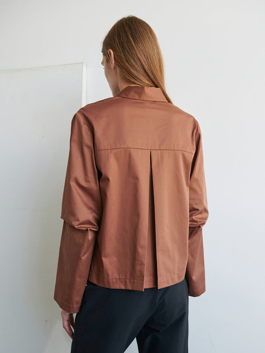 A person with long straight hair is standing with their back to the camera. They are wearing a Biker Shirt by Zero + Maria Cornejo, made from light brown cotton broadcloth featuring a pleat down the back, paired with black trousers. The background is plain and light-colored.