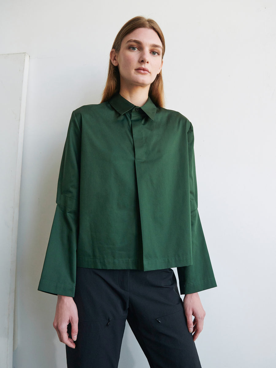 A person with long hair stands against a plain white background, wearing the Zero + Maria Cornejo Biker Shirt, a green cotton broadcloth, long-sleeved shirt with wide articulated sleeves, paired with black pants. Their expression is neutral, and their hands are relaxed by their sides.