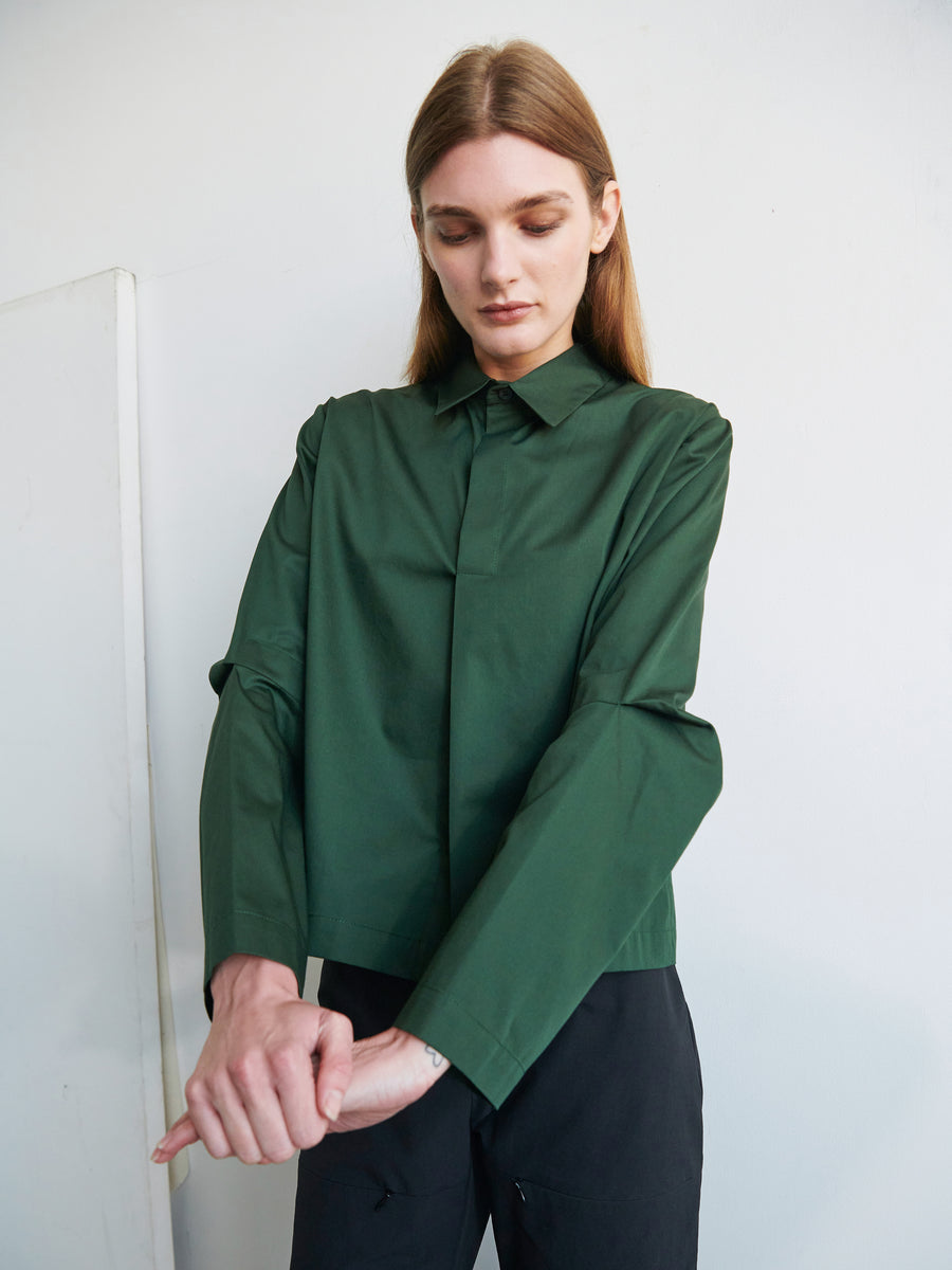 A person with long hair is standing against a plain background. They are wearing a dark green Biker Shirt from Zero + Maria Cornejo and black pants. The person is looking down and adjusting the articulated sleeve of their left arm with their right hand.