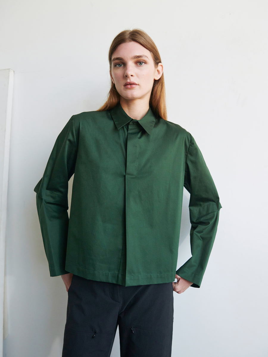 A person with straight, light brown hair stands against a plain background, wearing a dark green, long-sleeved Biker Shirt by Zero + Maria Cornejo and black pants. They have one hand on their hip while the other arm is relaxed by their side.