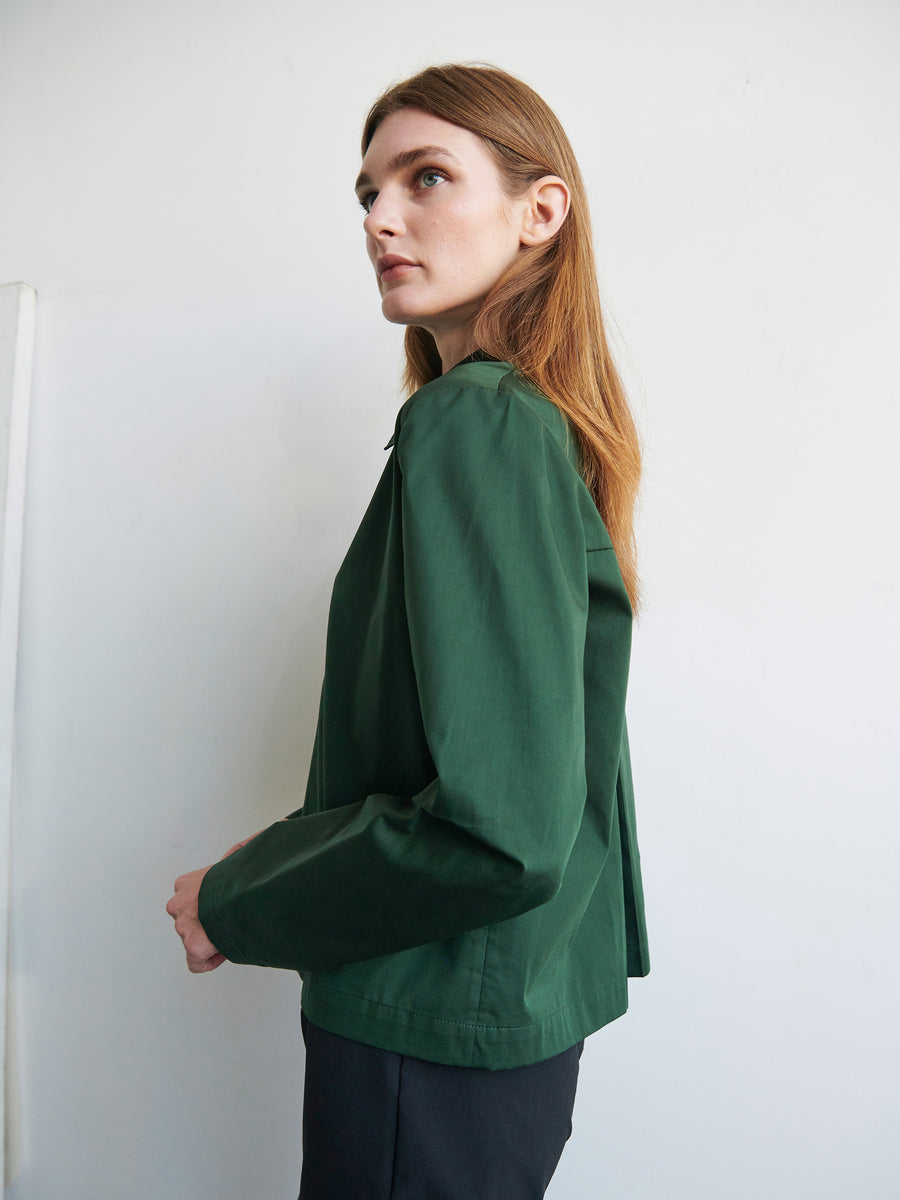 A person with long, light brown hair is standing against a light-colored background, looking to the side. They are wearing a forest green Biker Shirt by Zero + Maria Cornejo and dark pants. Their expression appears focused and thoughtful.