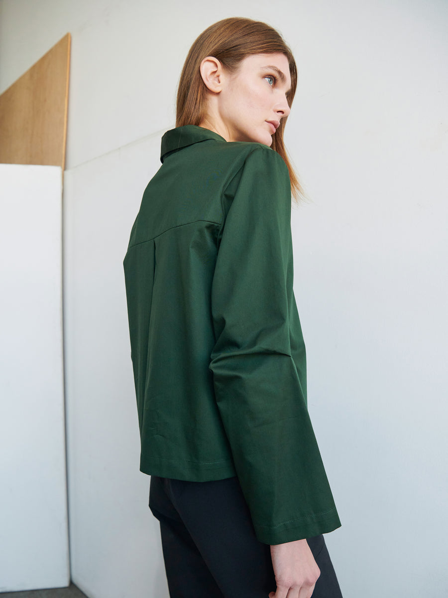 A person with long, light brown hair is seen from the side, looking away from the camera. They are wearing a dark green Zero + Maria Cornejo Biker Shirt made of cotton broadcloth and black pants. The background is plain and light-colored with a partial wooden panel on the wall.