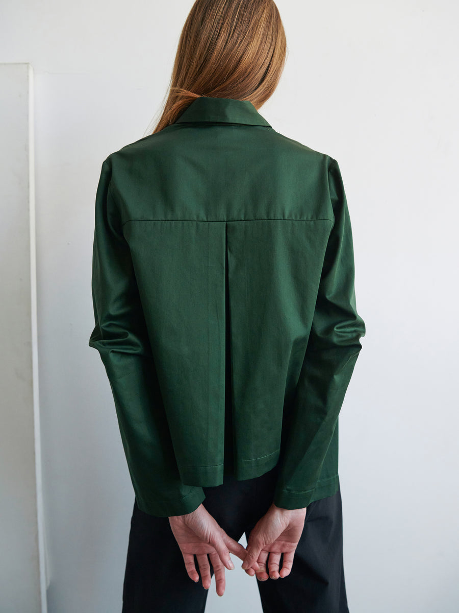 A person with long hair is standing with their back to the camera, wearing a dark green Zero + Maria Cornejo Biker Shirt with articulated sleeves and black pants. Their hands are clasped behind their back, and the background is a plain light-colored wall.