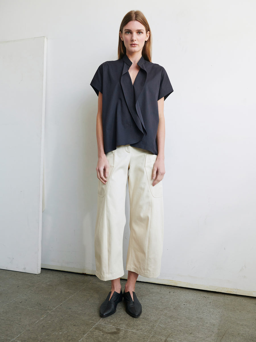 A person with long hair stands indoors against a plain white wall, wearing the Fin Gaban Shirt by Zero + Maria Cornejo, a short-sleeved, dark-colored top with a draped design and light-colored wide-leg pants, paired with black shoes. The floor is concrete, and a white panel leans against the wall.