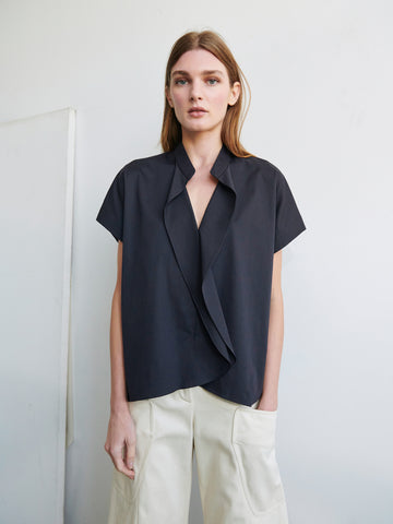 A woman with long brown hair stands against a plain white background. She is wearing a short-sleeved Fin Gaban Shirt by Zero + Maria Cornejo paired with light-colored pants, her hands casually in her pockets. She has a neutral expression and is looking directly at the camera.