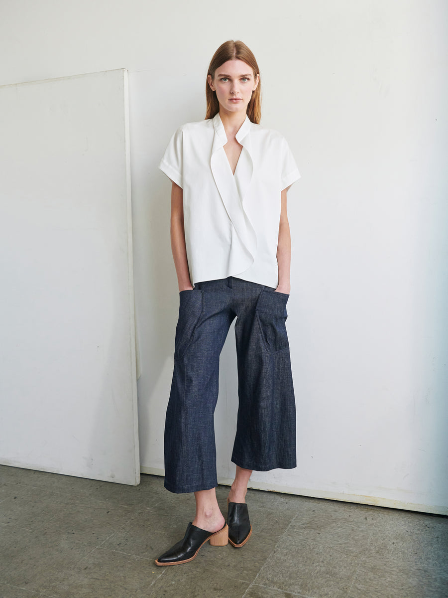 A person with straight brown hair is standing against a plain wall, wearing a Fin Gaban Shirt by Zero + Maria Cornejo, dark wide-leg cropped pants, and black mules with a wooden heel. Their hands are in their pockets, and they are looking directly at the camera.